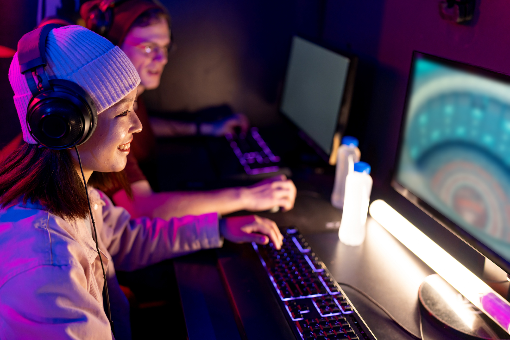 Two young adult gamers wearing headphones are concentrating on playing video games in a dark internet cafe, illuminated by colorful neon lights